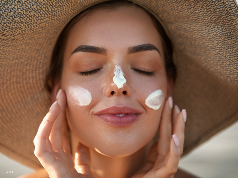 Photograph of a woman applying soothing cream sunscreen to face wearing a wide-brimmed hat, symbolizing effective sun protection and skin care.